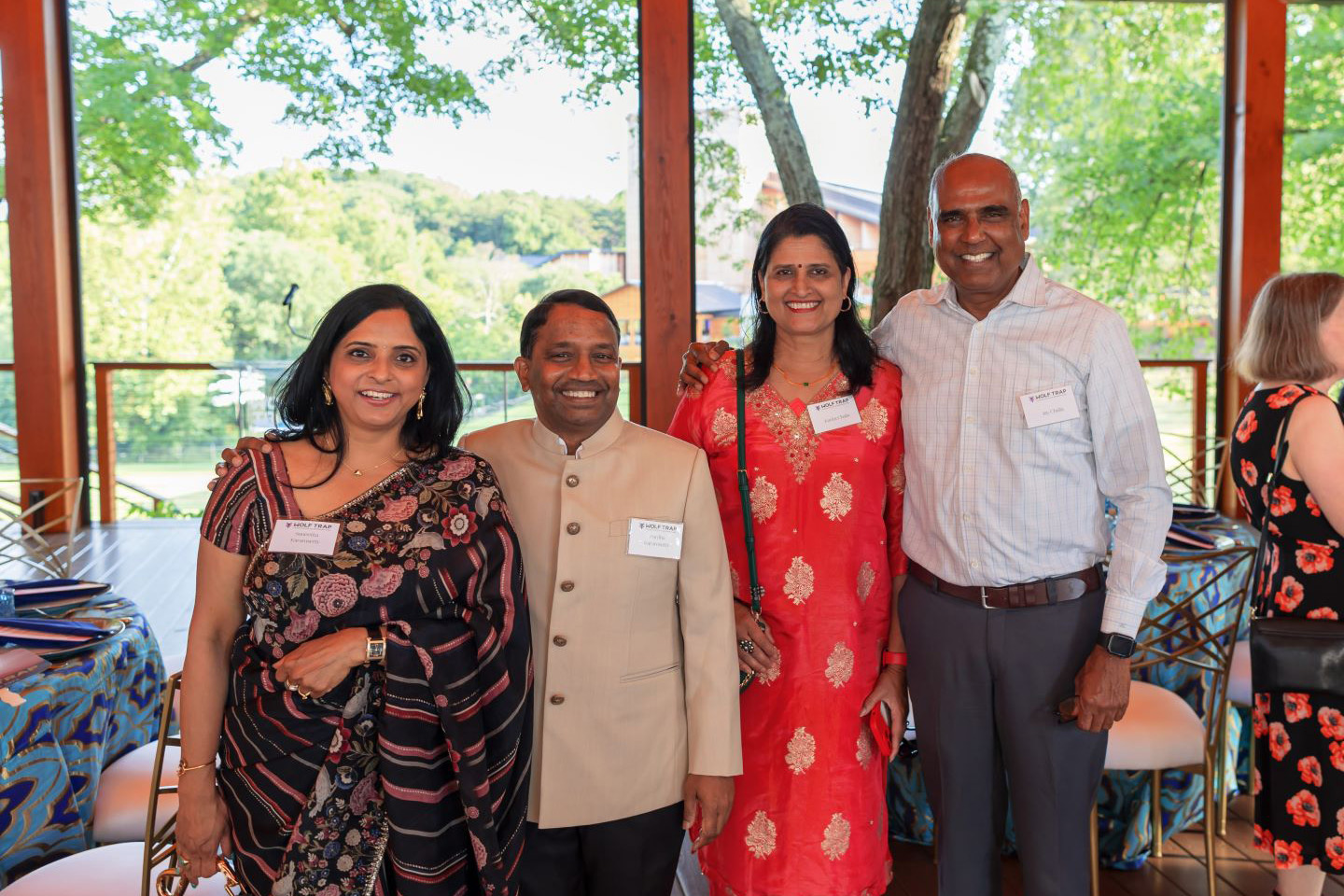 Dinner guests pose and smile on a Wolf Trap pavilion with green trees and the Filene Center in the background.