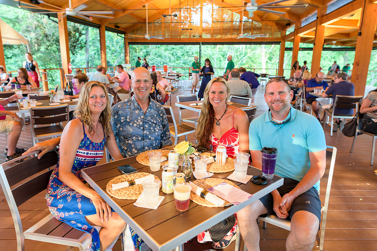 At a Wolf Trap Dinner and a Show event, four smiling guests at a table with trees in the distance.