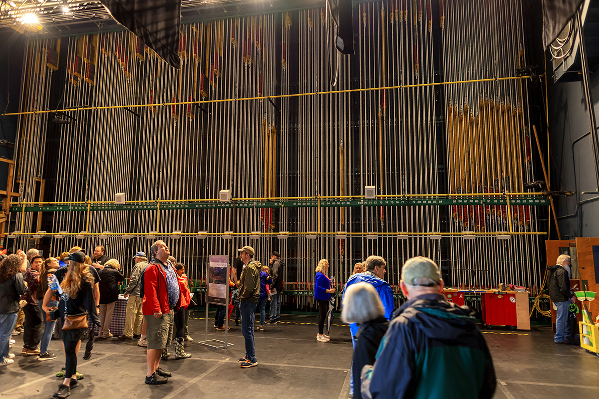 Wolf Trap members tour the Filene Center stage, with set rigging cables high in the background.