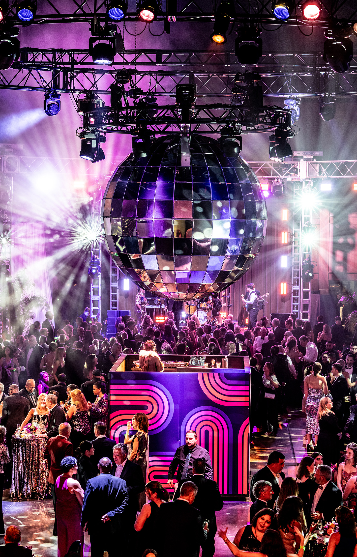 A dj spins at a colorful booth under a giant disco ball, surrounded by mingling guests.