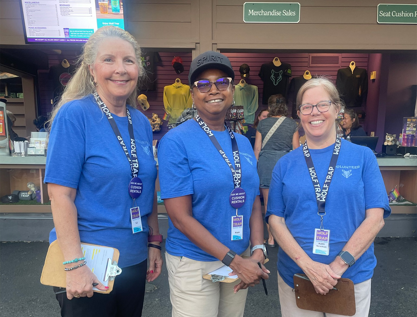 Three volunteers in blue shirts and lanyards smile in front of the Wolf Trap gift shop.