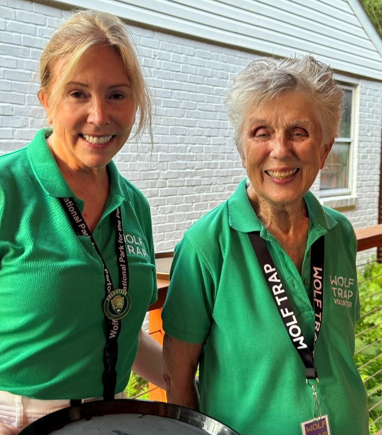 Bette Gorman and another volunteer smiling, wearing green shirts, holding serving trays.