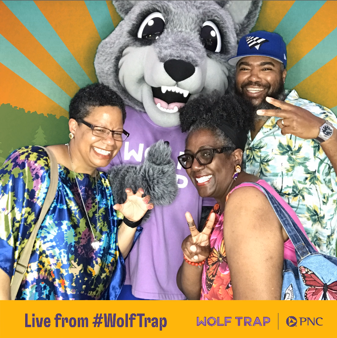 Three friends pose with Wolf Trap's mascot Wolfie, with a colorful background.