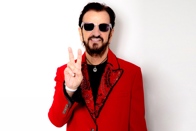 Ringo Starr in a red suit and sunglasses makes a peace sign.